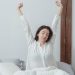 Young brunette with closed eyes sitting on comfortable bed under white blanket and stretching arms up while waking up in morning at home